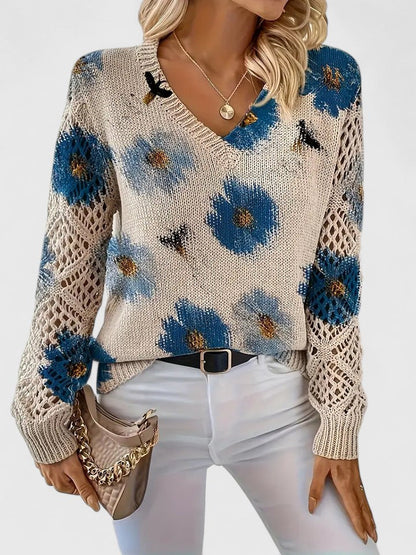 Frau trägt cremefarbenen Strickpullover mit blauen Blumen, V-Ausschnitt und Häkelärmeln, kombiniert mit weißer Hose und beiger Handtasche.
