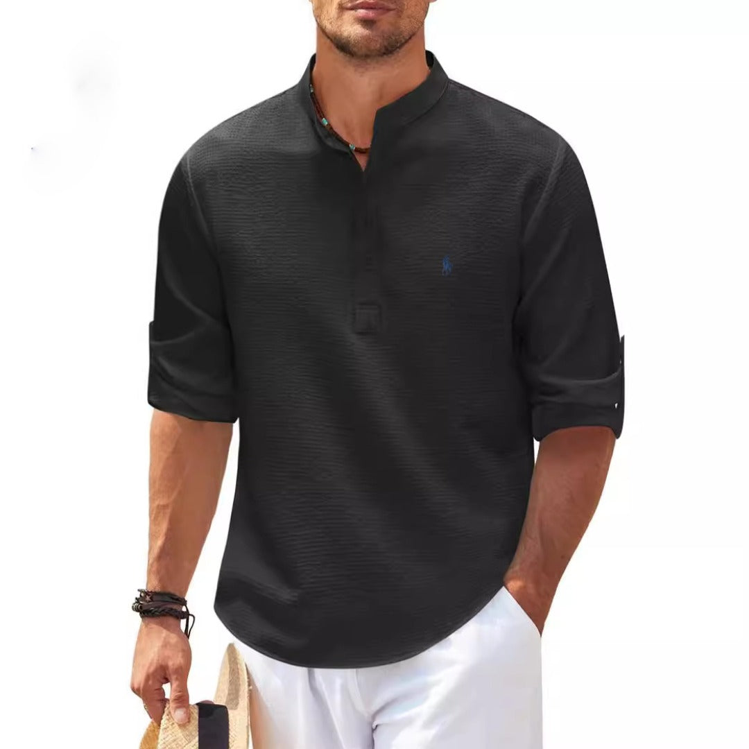 Mann in schwarzem, langärmligem Henley-Shirt mit Stehkragen, hält Strohhut, trägt weiße Hose. Freizeitmode, Herrenbekleidung, Sommeroutfit.