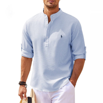 Mann in hellblauem Leinenhemd mit Stehkragen und hochgekrempelten Ärmeln, hält Strohhut. Sommermode, Freizeitlook, Herrenbekleidung.