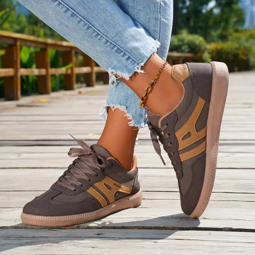 Damen Sneaker in Braun mit goldenen Streifen, getragen auf Holzsteg. Modische Freizeitschuhe, ideal für Herbstmode und Outdoor-Aktivitäten.