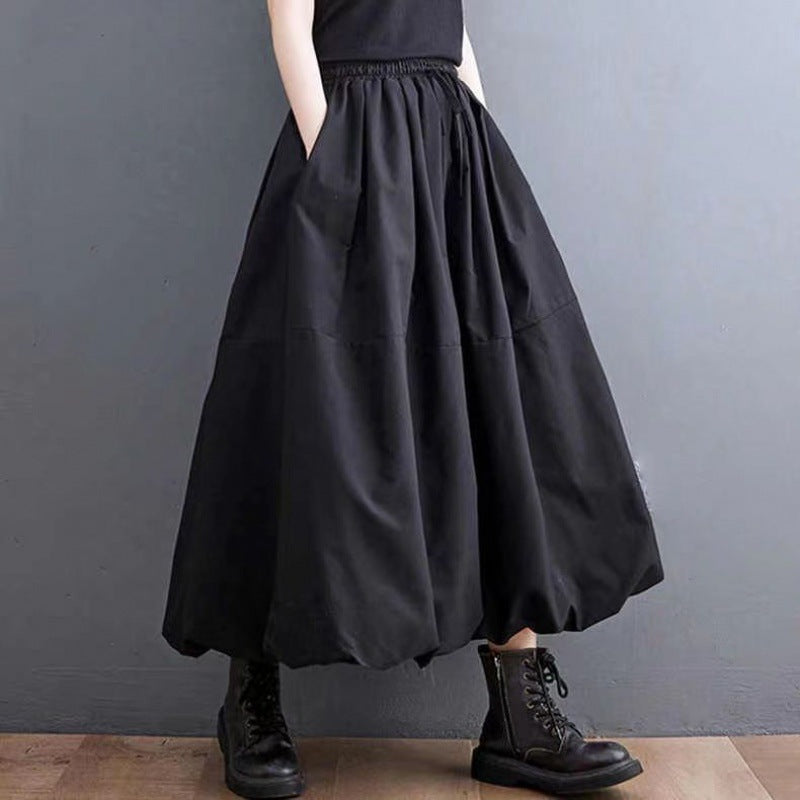 Person in schwarzem, langem Rock mit Taschen und schwarzen Stiefeln vor grauer Wand. Modetrend, Gothic-Stil, lässiges Outfit.
