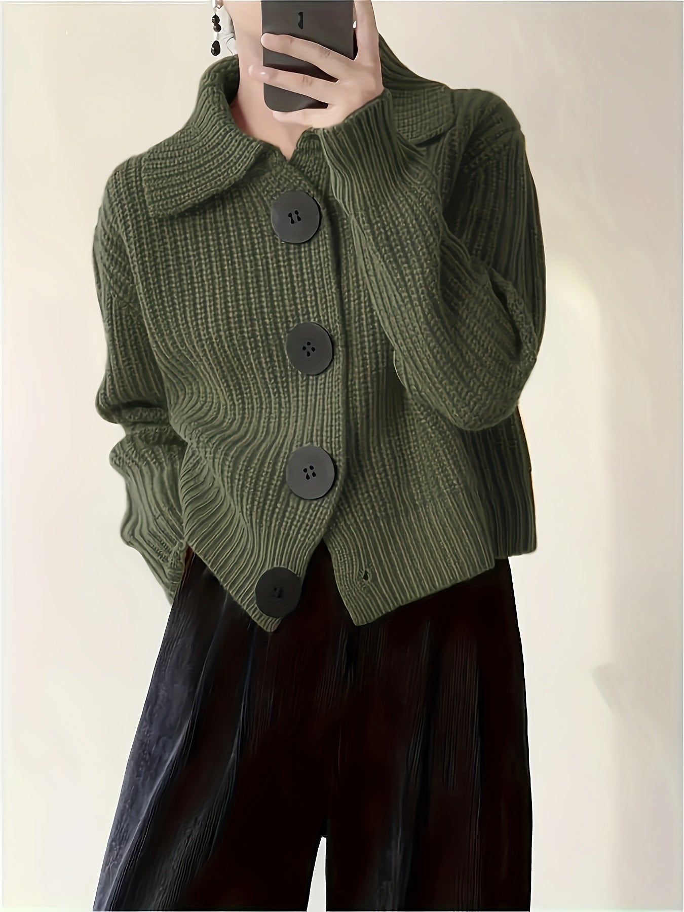 Dunkelgrüne Strickjacke mit großen schwarzen Knöpfen, getragen von einer Person, die ein Smartphone hält. Modetrend, Herbstmode, Damenbekleidung.