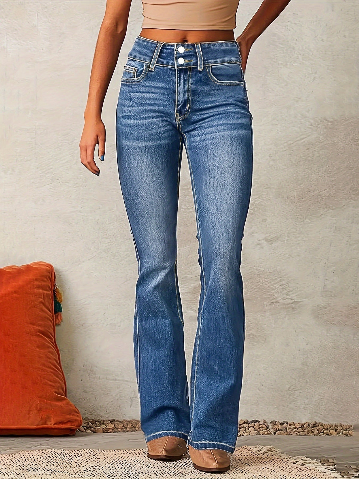Frau in blauen Schlagjeans und beigem Top, stehend vor einer neutralen Wand. Modetrend, Damenjeans, lässiger Stil, bequeme Passform.