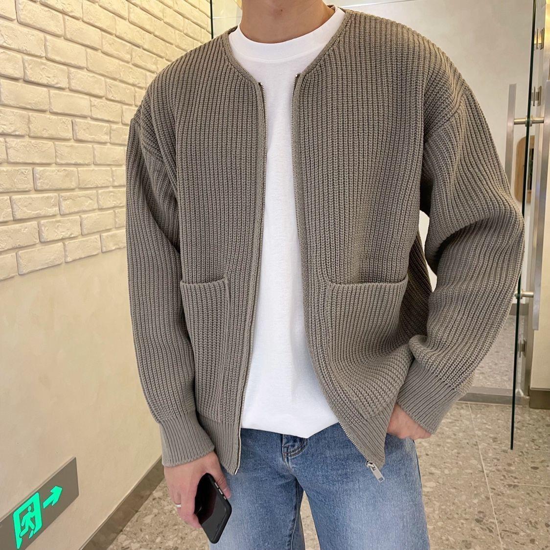 Mann in grauer Strickjacke und weißem T-Shirt, hält Smartphone, steht vor Ziegelwand. Modetrend, lässiger Herrenstil, Herbstmode.