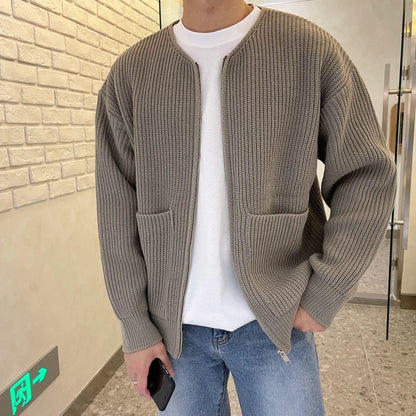 Mann in grauer Strickjacke und weißem T-Shirt, hält Smartphone, steht vor Ziegelwand. Modetrend, lässiger Herrenstil, Herbstmode.