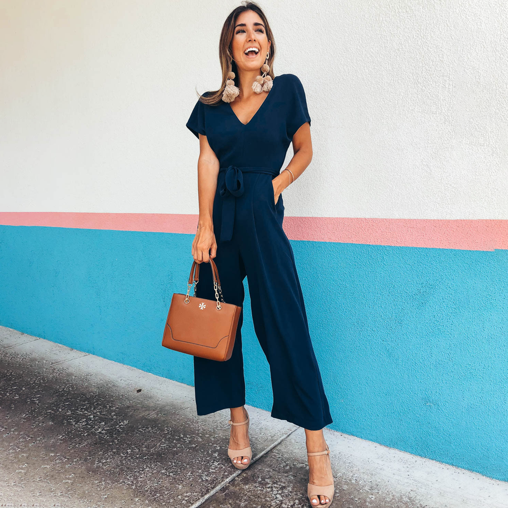 Frau in dunkelblauem Jumpsuit mit brauner Handtasche vor bunter Wand. Modetrend, Sommeroutfit, elegante Damenmode, stilvolle Accessoires.