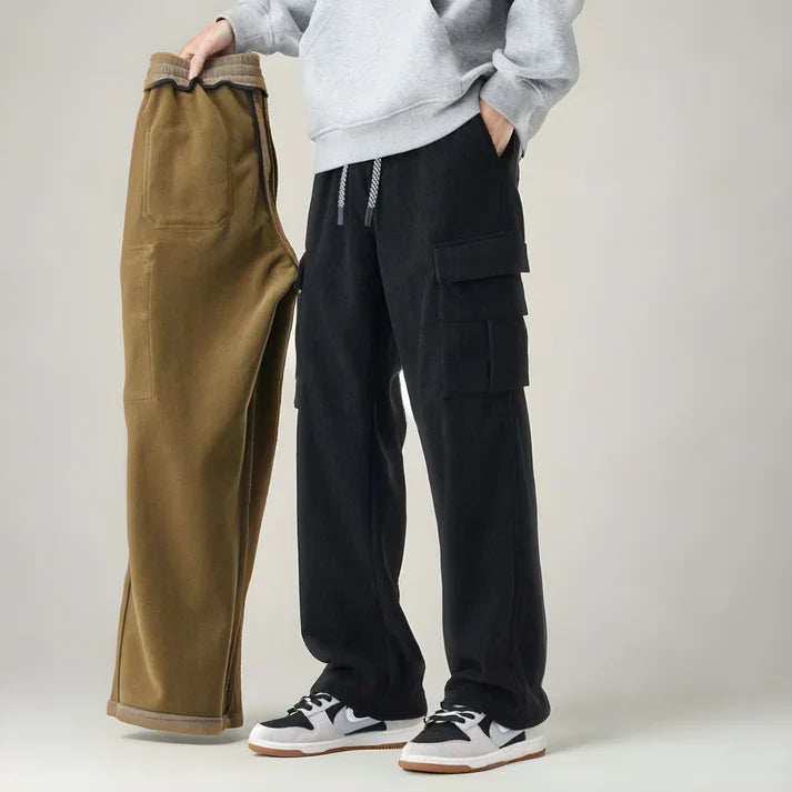 Person in grauem Hoodie hält braune Hose, trägt schwarze Cargohose und weiße Sneakers. Modetrend, lässiger Stil, Herrenbekleidung.