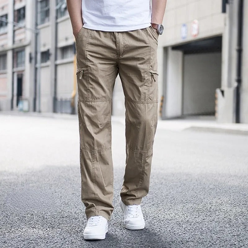 Mann in beigen Cargohosen und weißen Sneakers steht auf städtischer Straße. Moderne Herrenmode, lässiger Stil, urbane Umgebung.