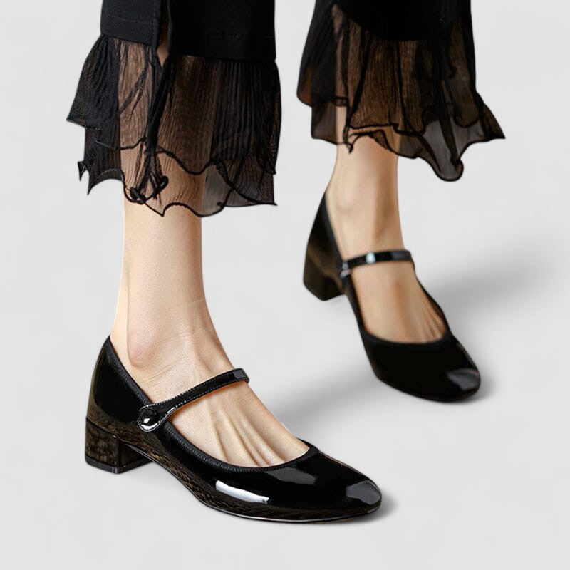 Schwarze Lackleder-Mary-Jane-Schuhe mit niedrigem Absatz, getragen von einer Person in schwarzer, transparenter Rüschenhose. Elegante Damenmode.