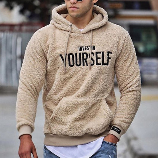Mann in beigem Fleece-Hoodie mit Aufschrift "Invest in Yourself", lässiger Stil, Jeans, urbaner Hintergrund. Modetrend, Herrenbekleidung, Streetwear.