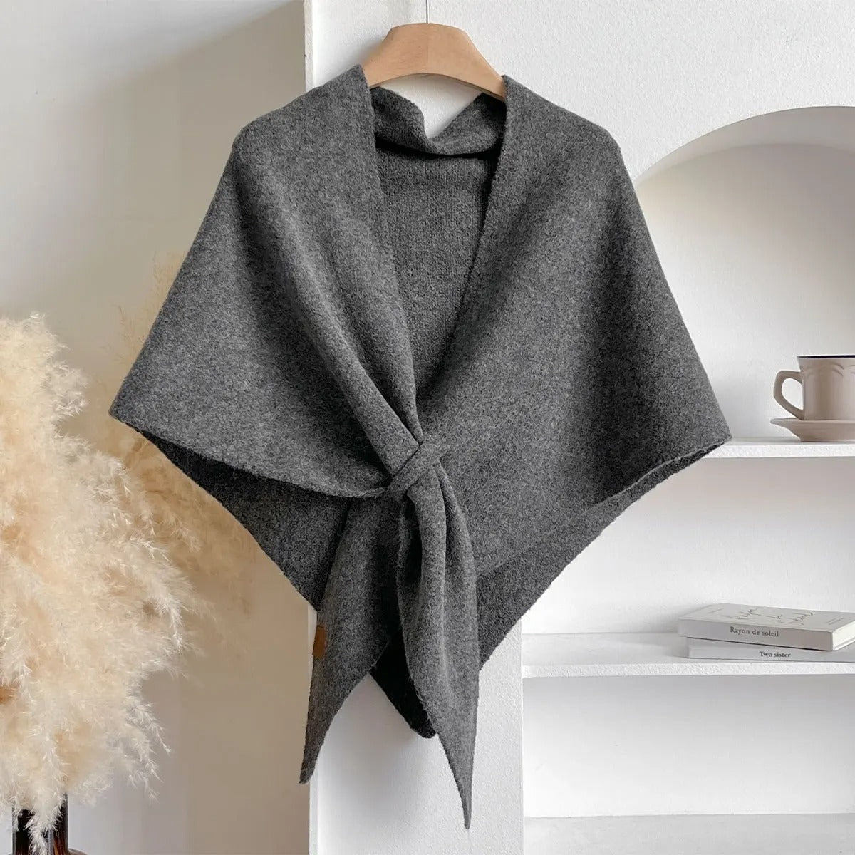 Grauer Wollschal auf Holzkleiderbügel, drapiert vor weißer Wand. Elegantes, warmes Accessoire für Herbst und Winter. Ideal für modische Outfits.