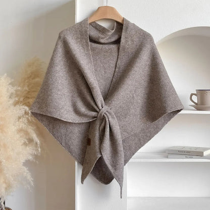 Grauer Wollponcho auf Holzkleiderbügel, drapiert vor weißem Regal mit Tasse und Büchern. Elegante Herbstmode, gemütlicher Strick, stilvolles Accessoire.