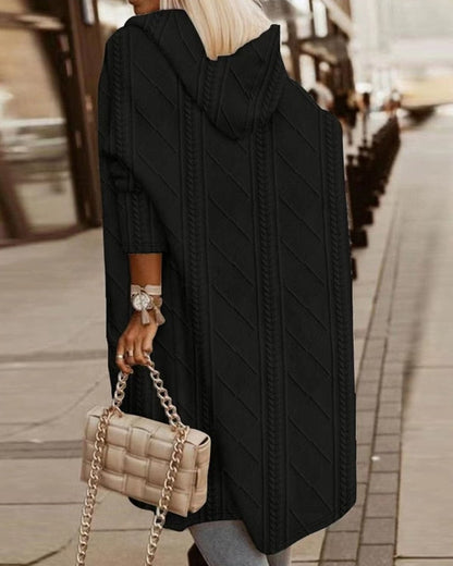 Frau in schwarzem Strickmantel mit Kapuze, trägt beige gesteppte Handtasche mit Kettengriff, geht auf städtischem Gehweg.