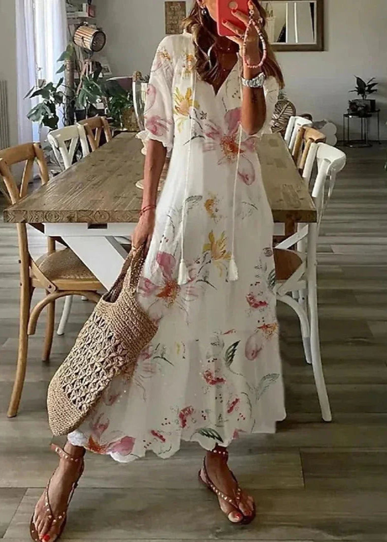 Frau in geblümtem Sommerkleid, hält Strohtasche, steht in modernem Esszimmer mit Holztisch und Stühlen. Perfektes Outfit für warme Tage.